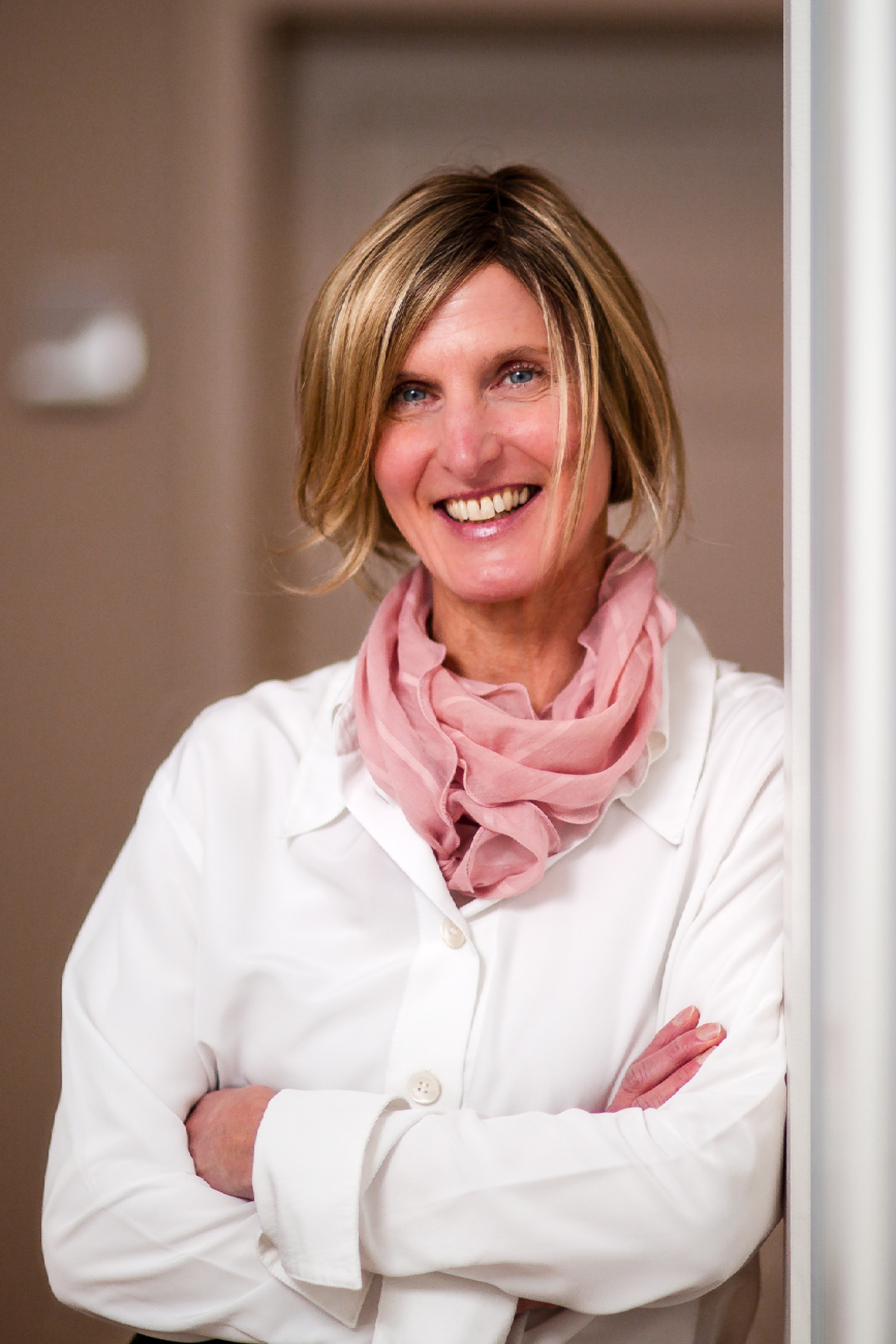 Eine lächelnde Frau mit blonden, kinnlangen Haaren steht in einem hellen Flur, sie trägt einen weißen Kittel und einen rosafarbenen Schal. Ihre Arme sind locker verschränkt.
