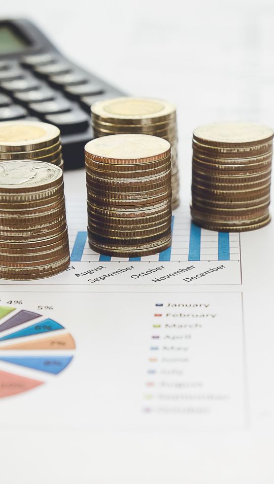 Gestapelte Münzen stehen auf einem Blatt mit Diagrammen und Tabellen. Daneben liegt ein Taschenrechner.